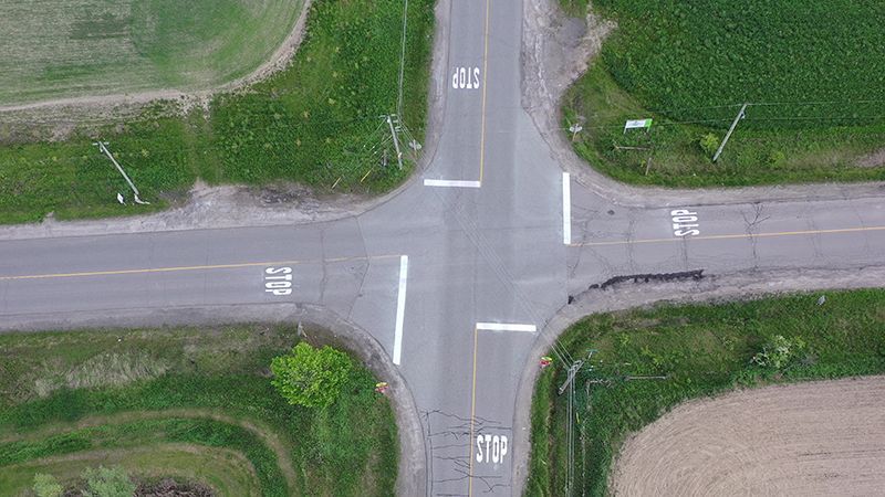 Overhead view of a rural, four-way intersection with stop signs, white road markings, and green fields.