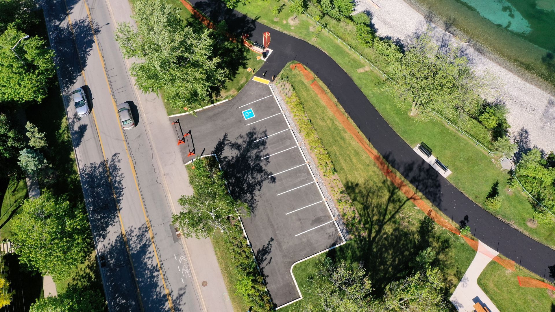 Overhead view of a parking lot next to a path and road, near a lake. Includes accessible parking.