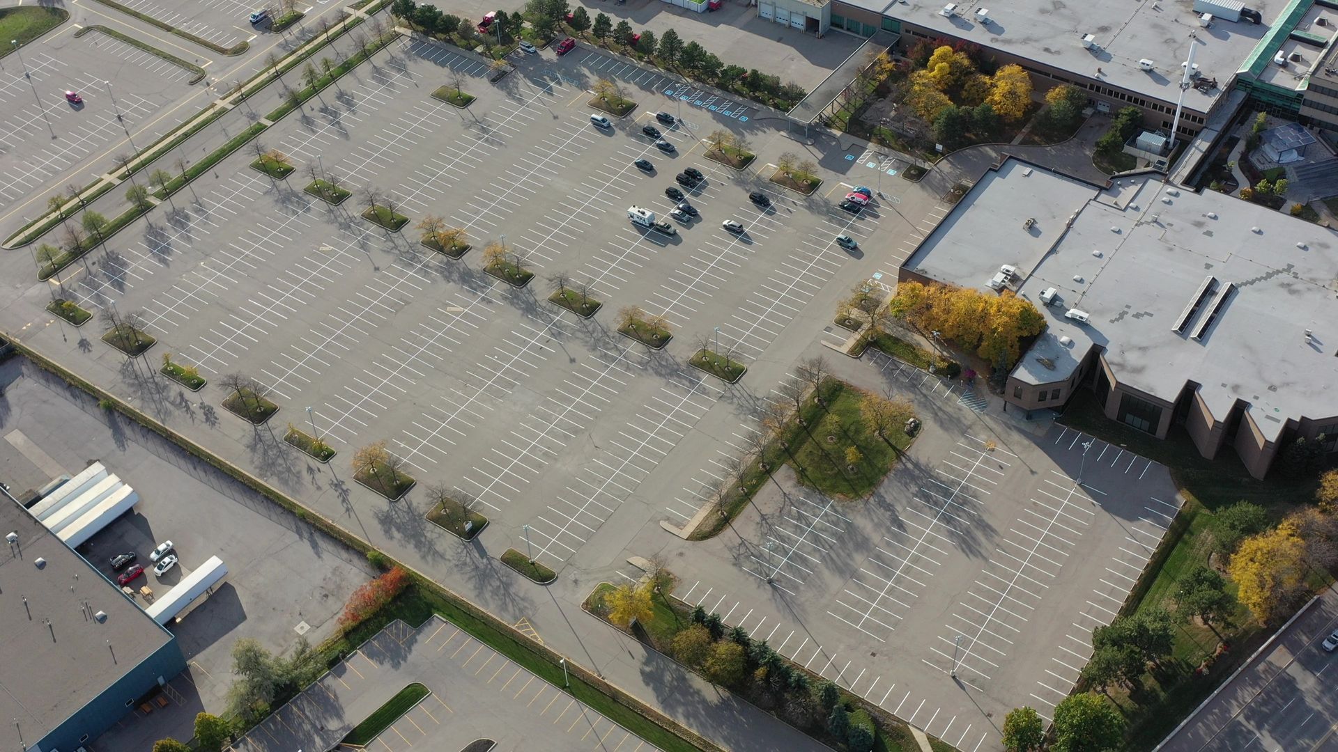 Aerial view of a mostly empty parking lot with some cars parked. Buildings and trees surround the lot.