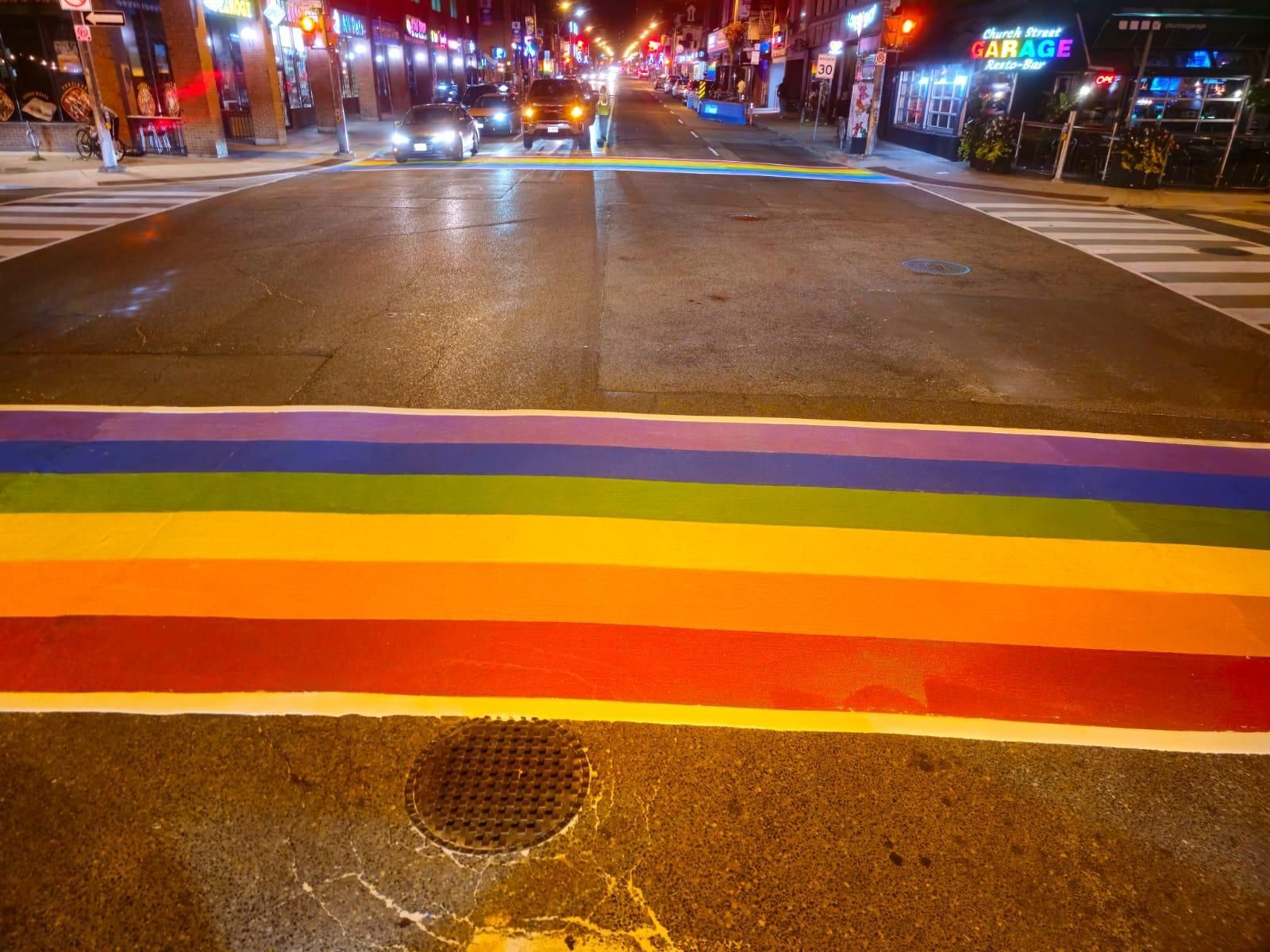 Rainbow crosswalk in city street at night, cars approaching.