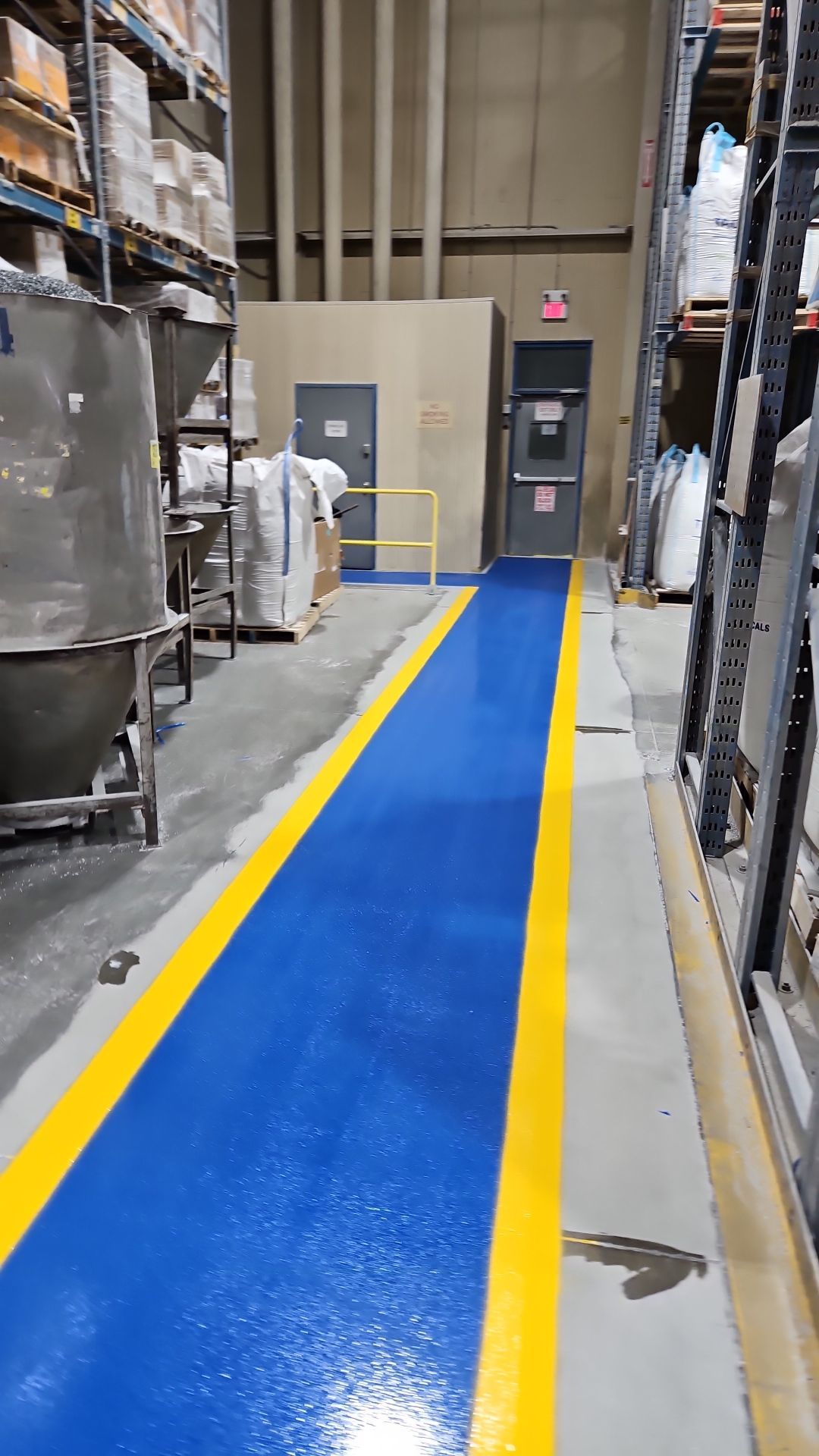 Warehouse aisle with blue and yellow painted floor, storage racks, and doors.