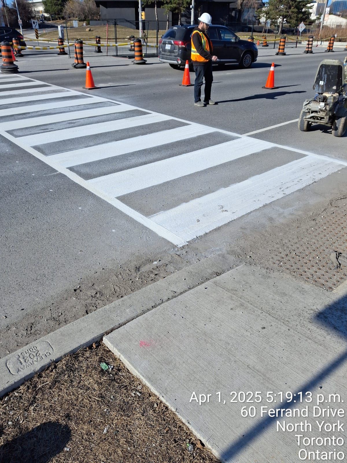 Crosswalk being painted; worker in safety vest, orange cones, car passing. North York, Ontario.