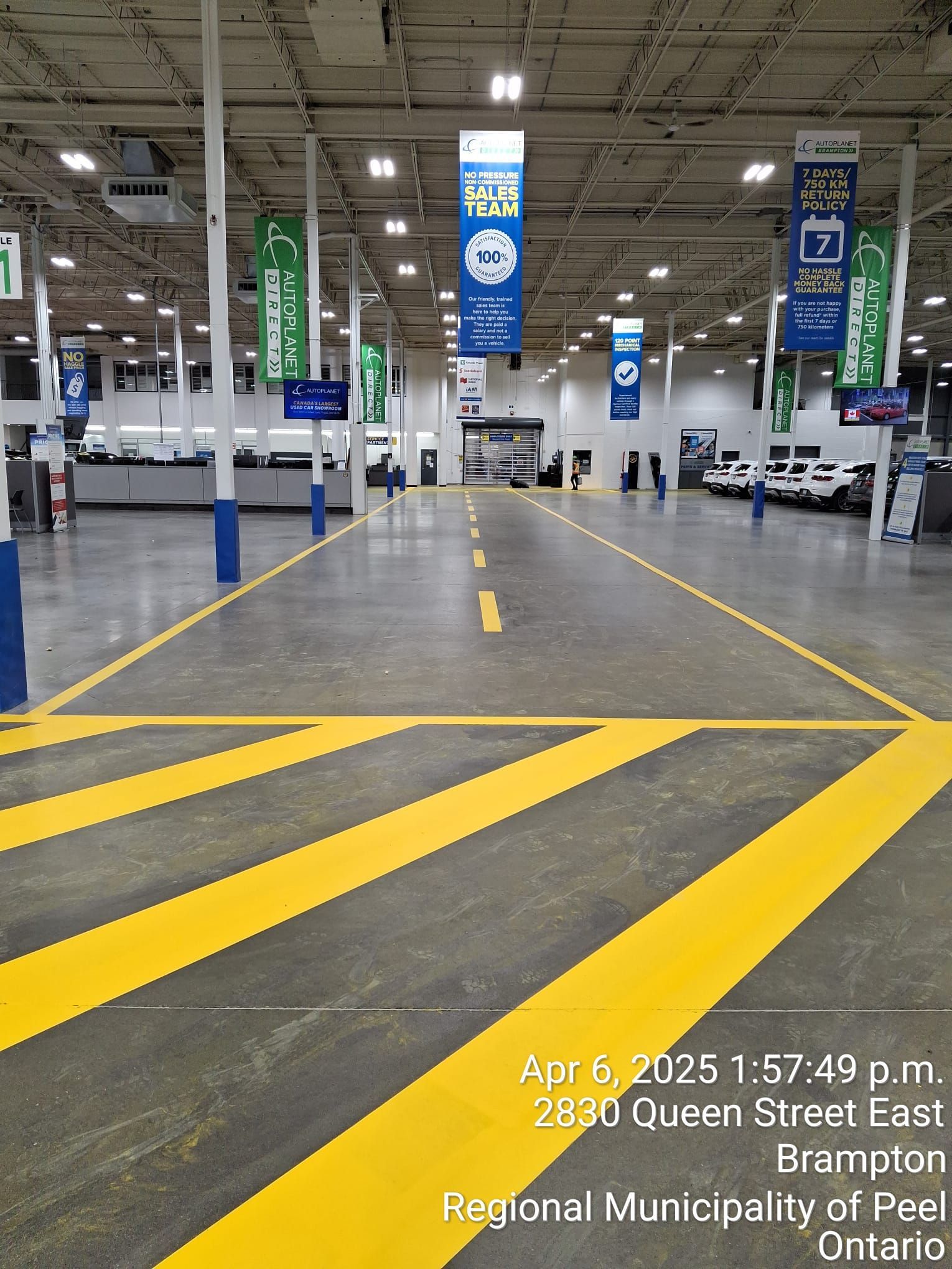 Interior view of an empty warehouse-like space, yellow lane markings, blue support beams, banners, text: 2830 Queen Street East, Brampton.