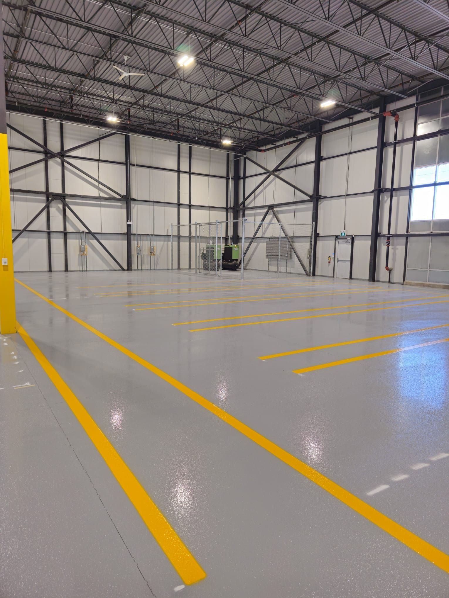 Empty indoor space with gray floor and yellow markings; metal framework and high ceiling.