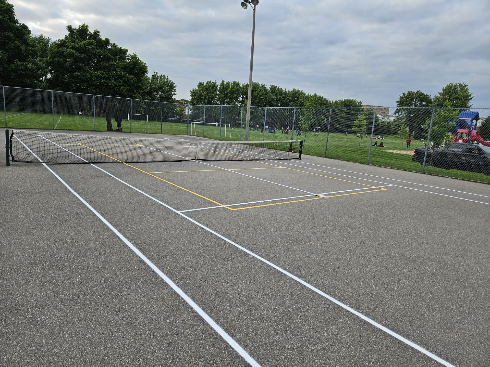 Asphalt sport court with painted white and yellow lines, chain link fence, and a light pole.