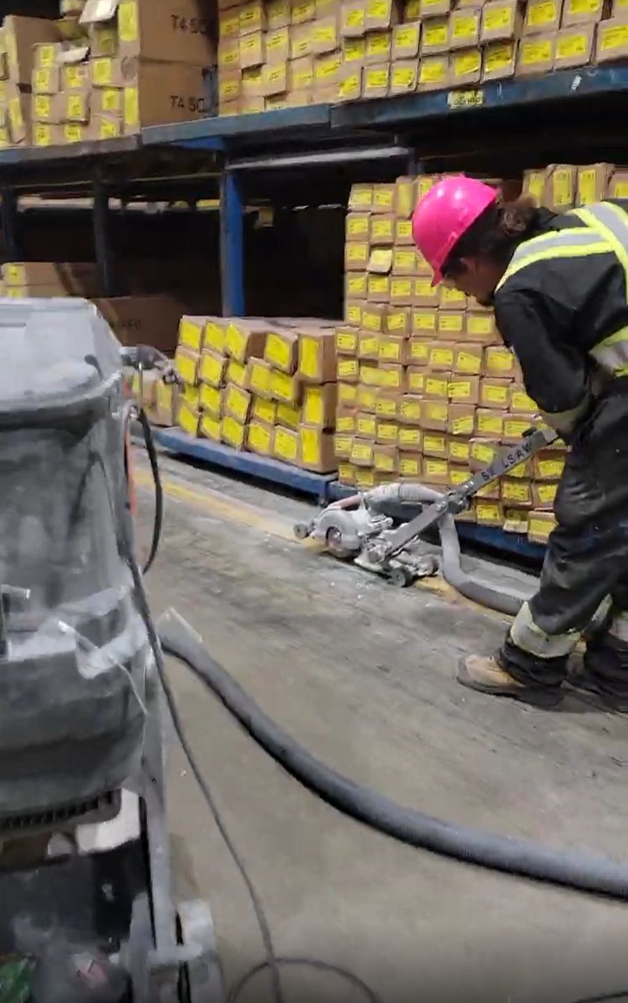 Person in pink hard hat using a hose with a machine, near boxes on shelves.