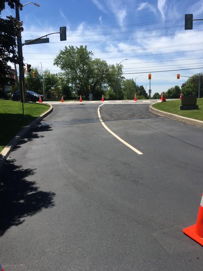 Road with fresh asphalt, orange cones, traffic lights, and a tree under a blue sky.
