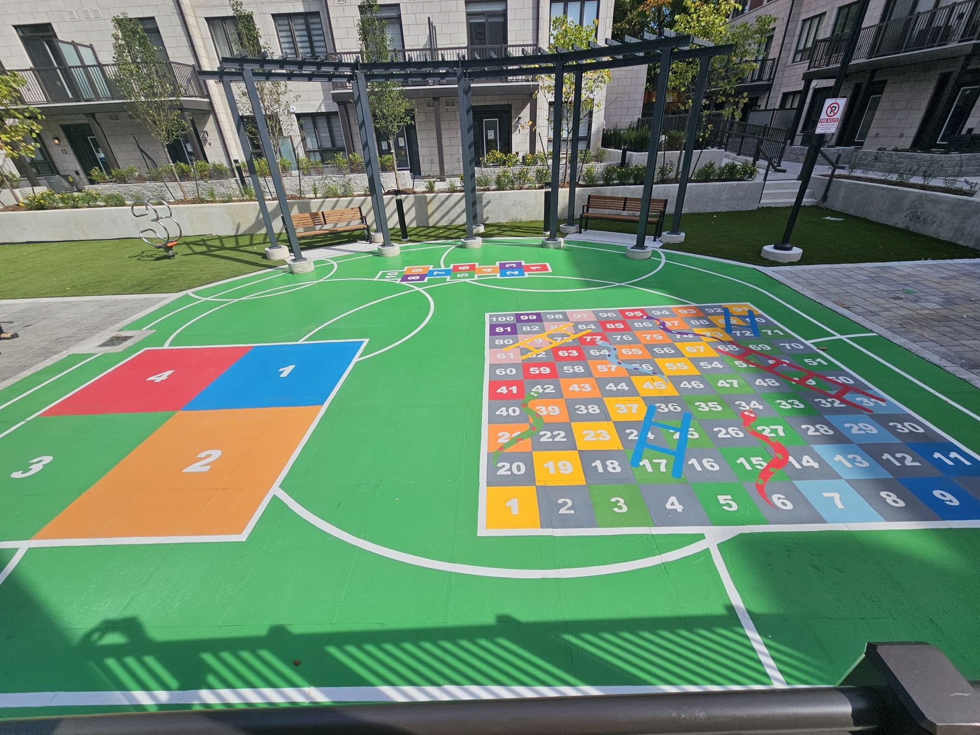 Green outdoor play area with painted games, a pergola, and surrounding buildings.