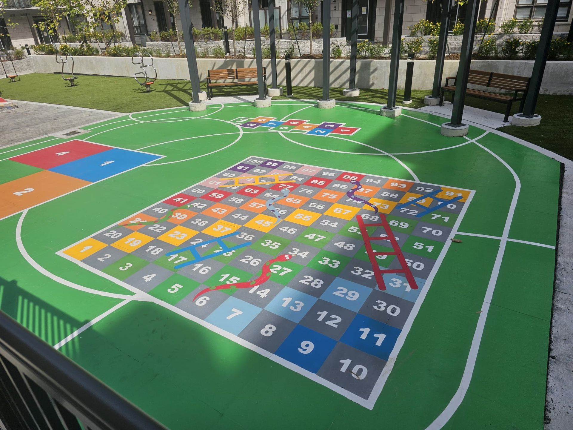 Outdoor Snakes and Ladders game painted on a green surface, with basketball court lines, surrounded by a grassy area.