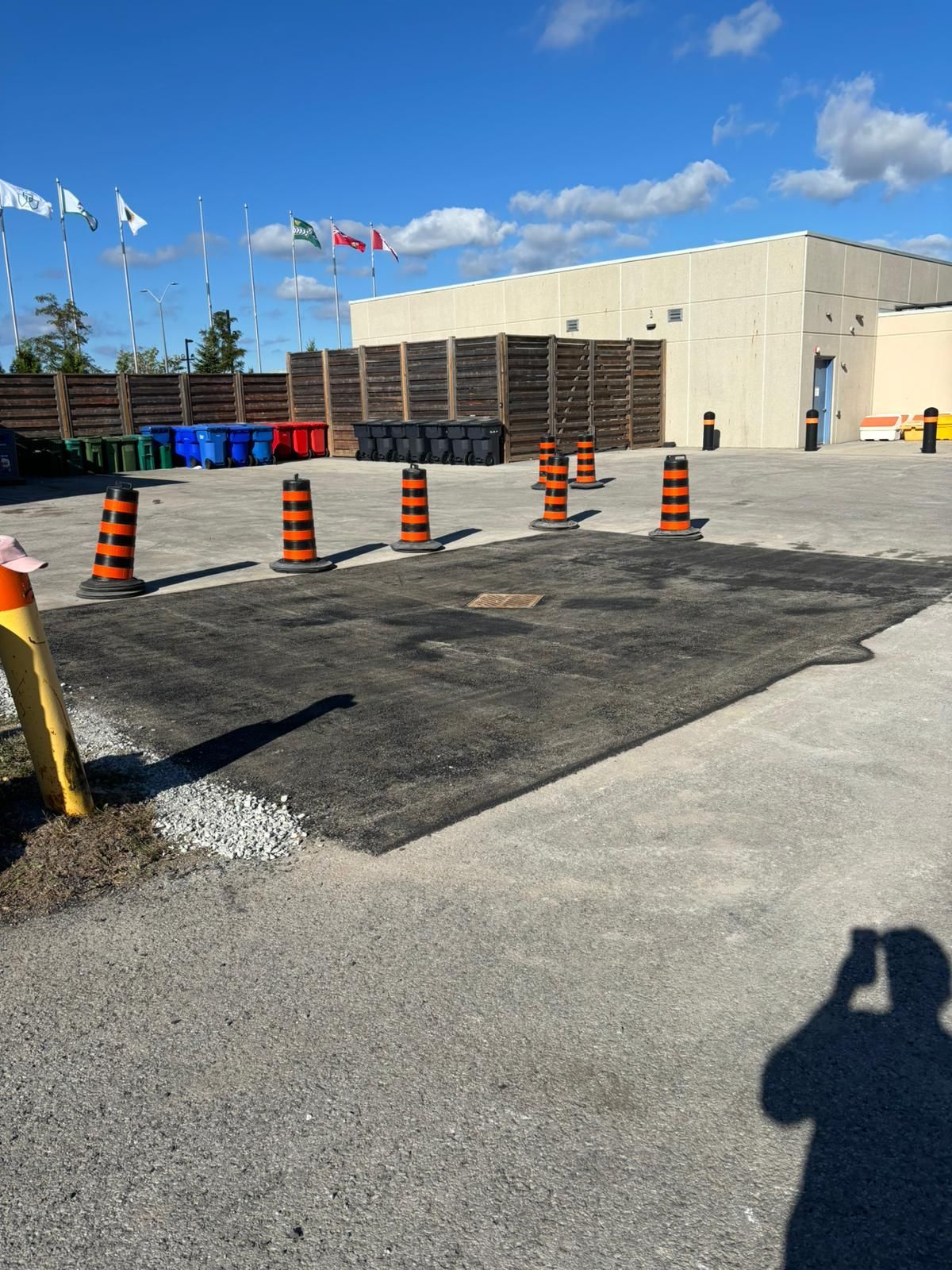Asphalt repair in parking lot, orange traffic cones. Recycle bins and building in background. Sunny day.