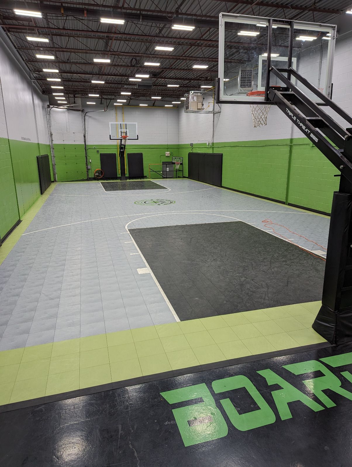 Indoor basketball court with green and gray floor tiles, black backboards, and fluorescent lighting.