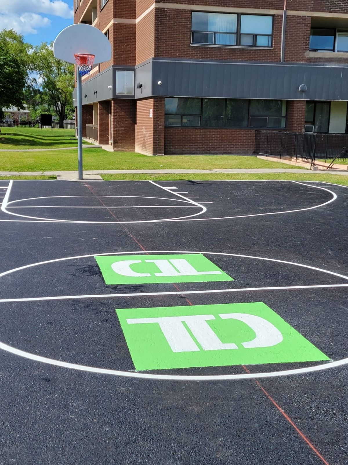 Basketball court with painted lines and green logo squares, in front of a brick building.