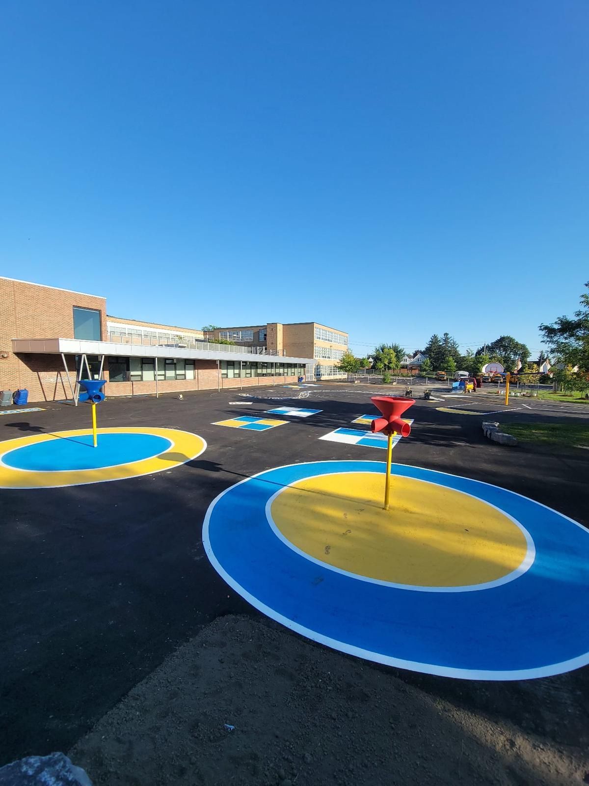 Playground with blue and yellow circles, water features, and a school building under a clear blue sky.