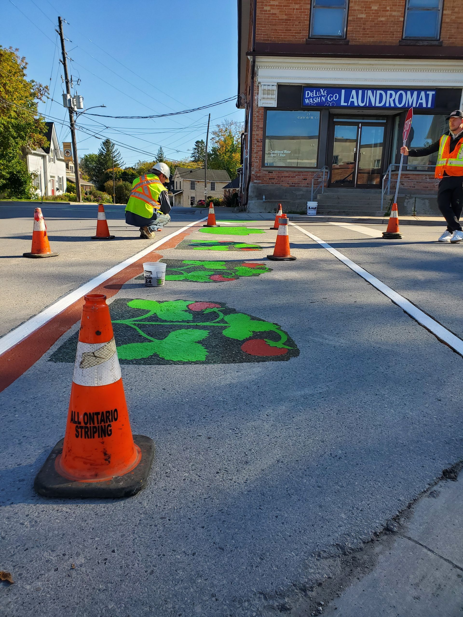 Workers painting colorful art on a crosswalk, marked by orange cones. Exterior daytime shot in front of a brick building.