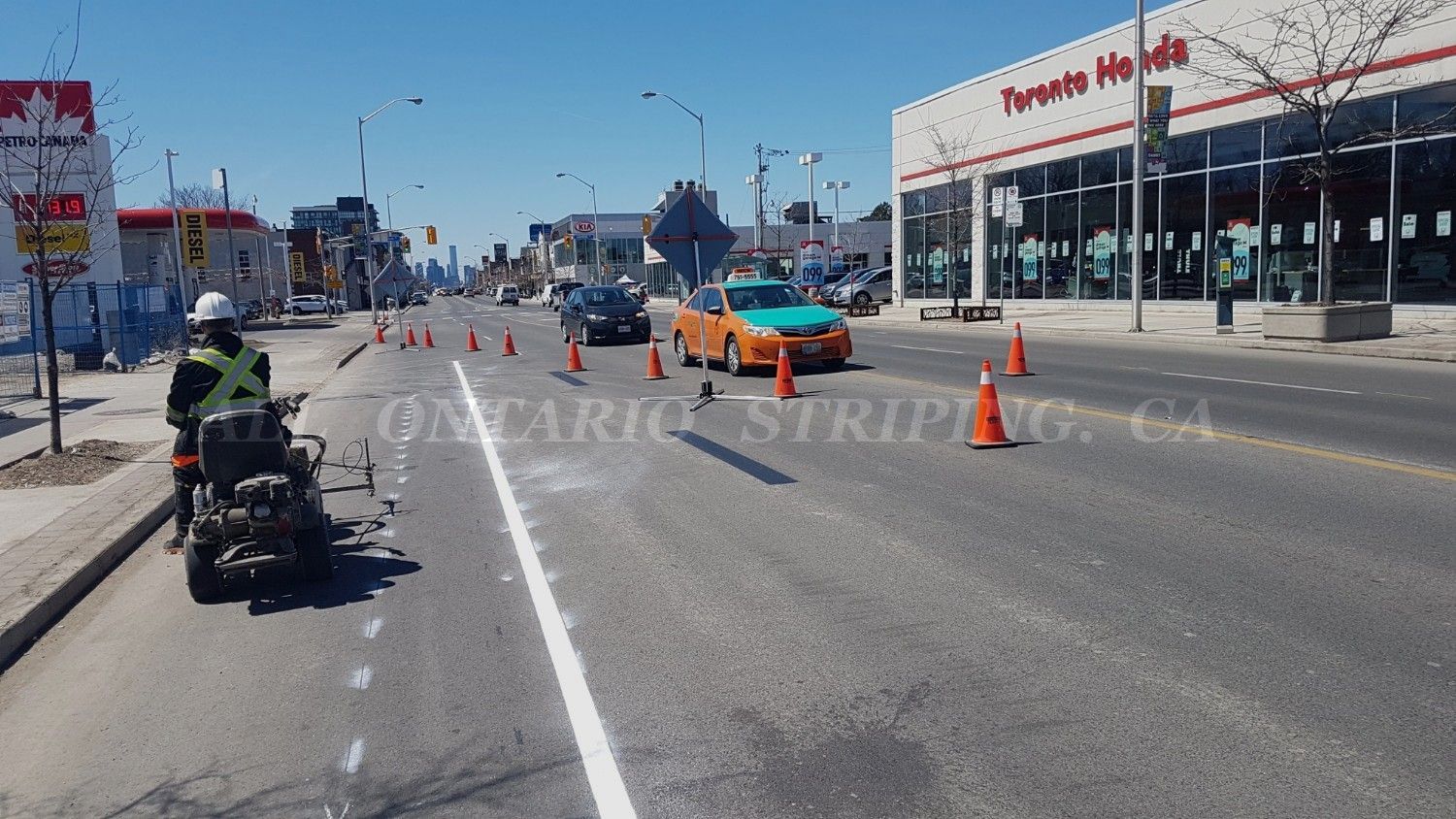 Road worker striping a street; orange cones, cars, and a Toyota dealership visible.