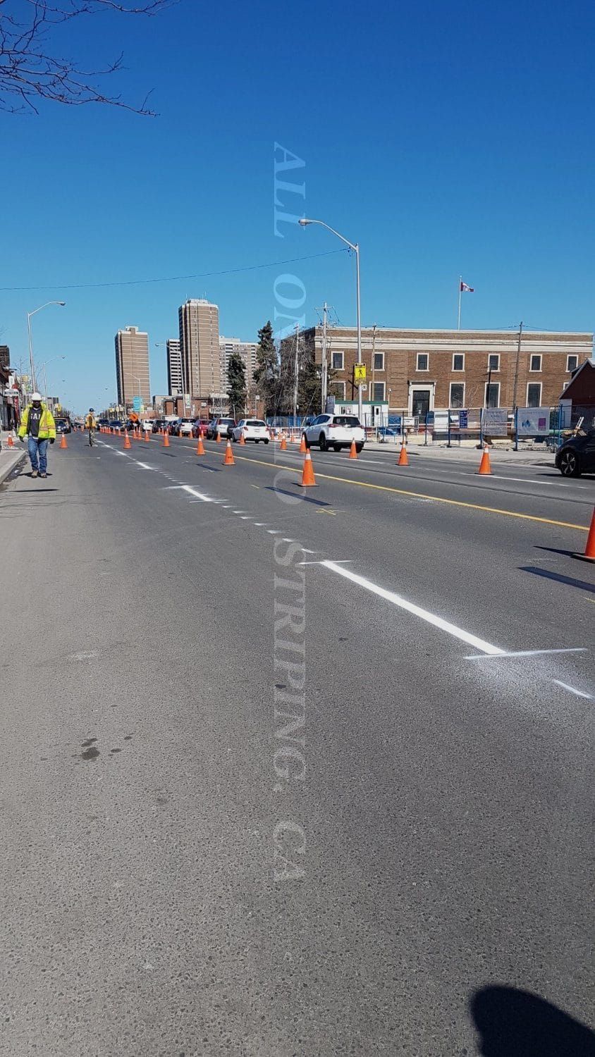 Road with orange cones and white lines, possibly road work. Buildings in the background on a sunny day.