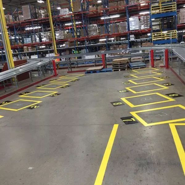 Warehouse floor with yellow painted boxes for pallet placement. Conveyor belts and shelves visible.