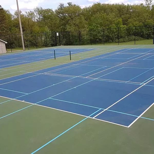Blue and green outdoor sports courts, likely for tennis or pickleball. Fenced area with trees in the background.