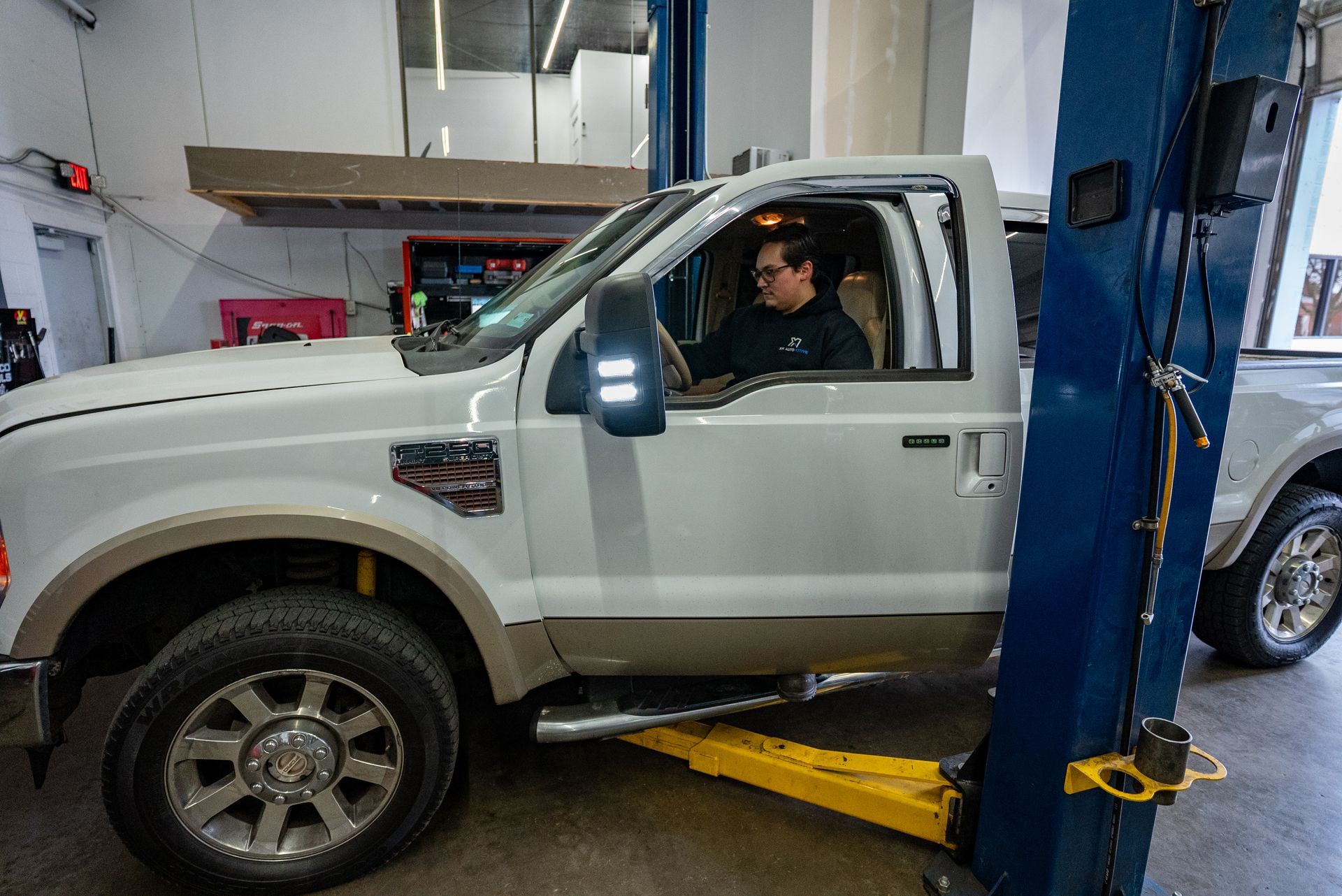 White and tan pickup truck on a lift inside a repair shop, driver inside | Lenexa, KS