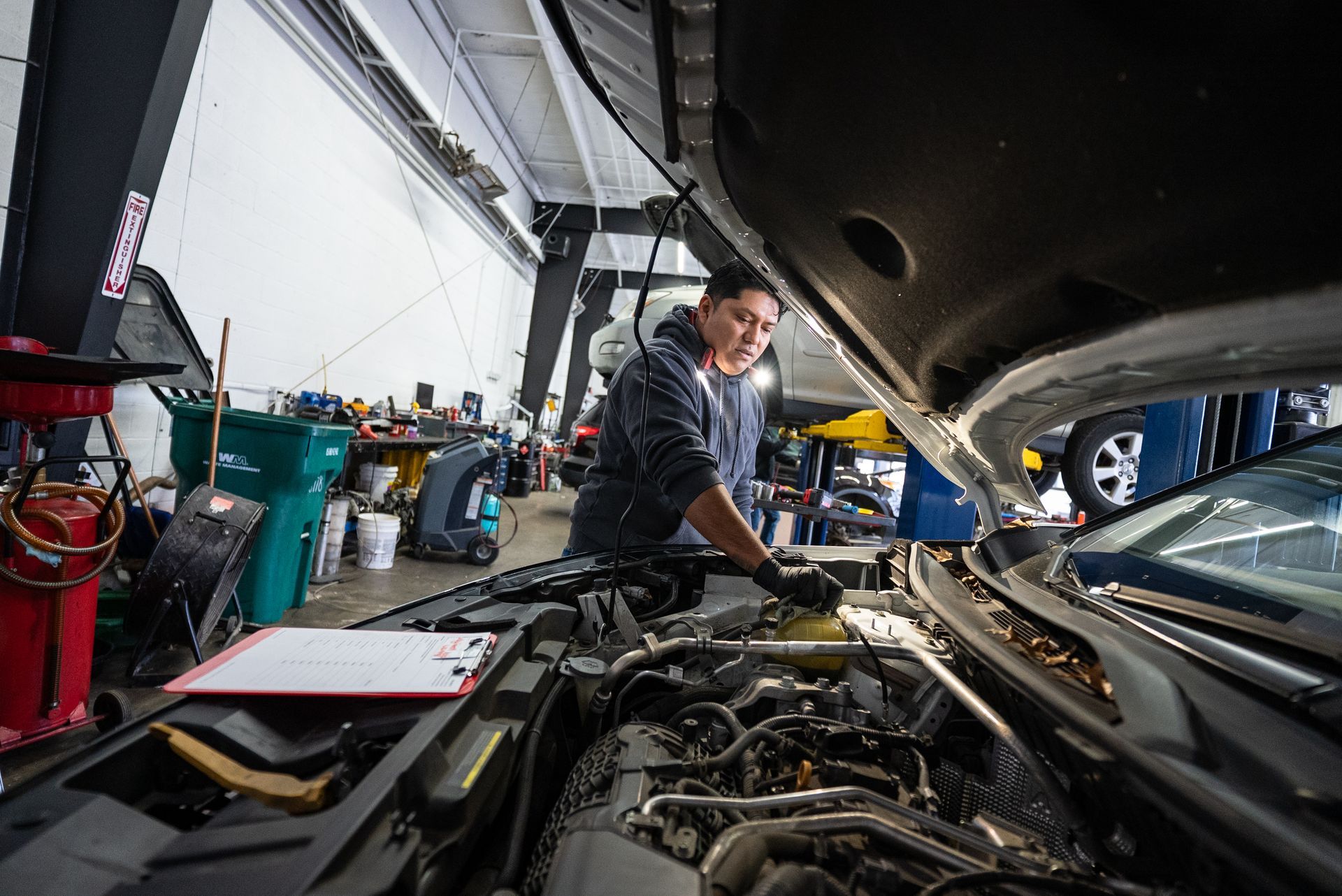 Mechanic inspecting a car engine in a well-lit auto shop, wearing gloves, with the hood open | Lenexa, KS