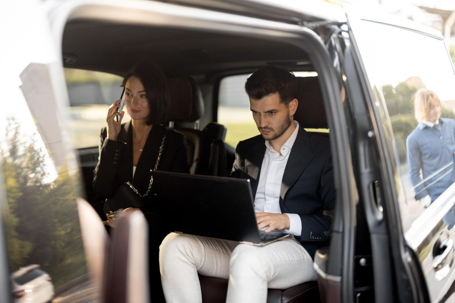 A man and a woman are sitting in the back seat of a car using laptops.