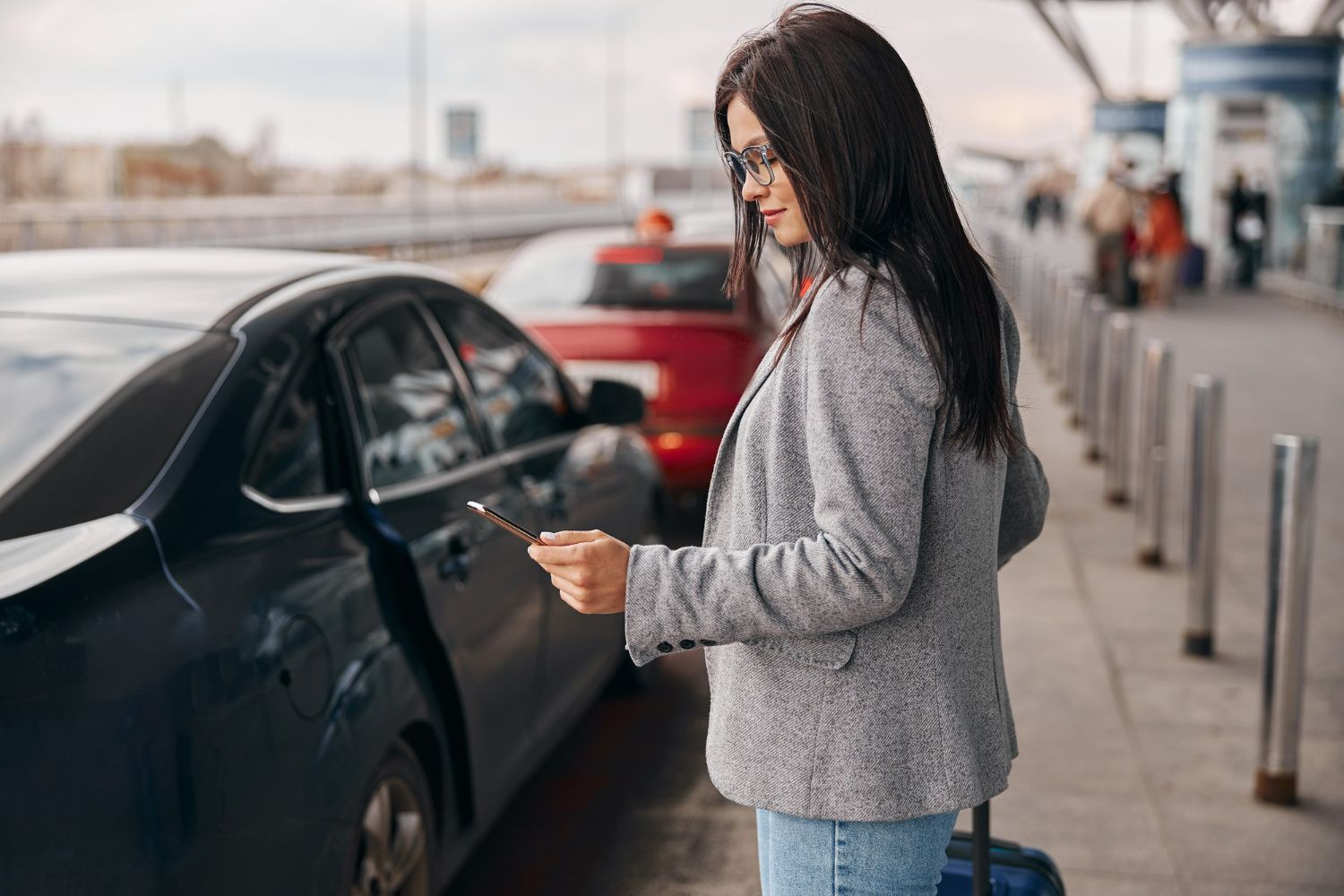 A woman is standing next to a car at an airport looking at her phone.