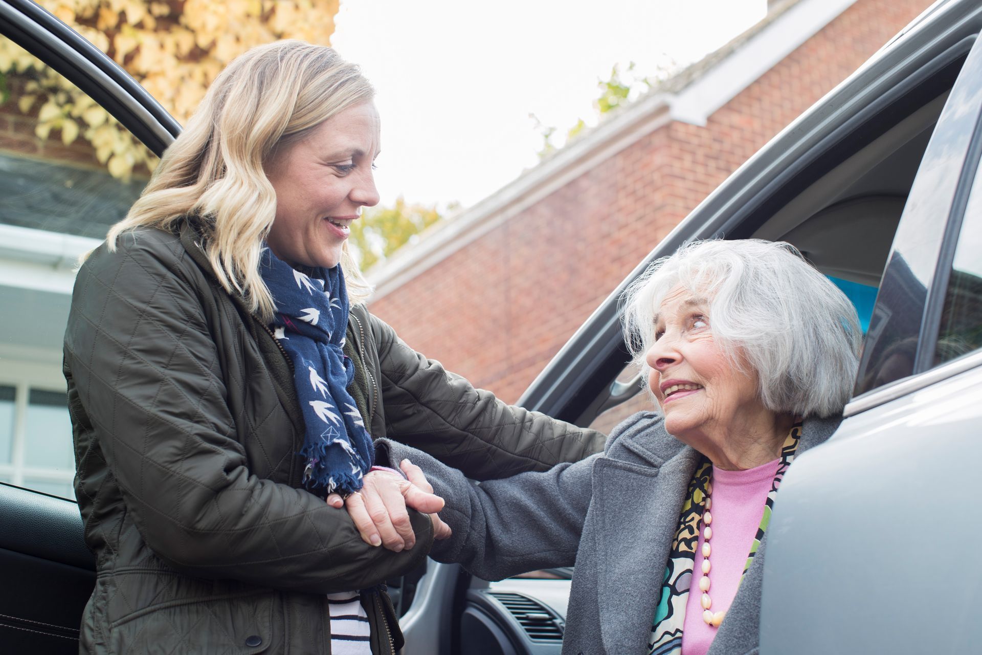Woman helps elderly person exit a car; both smile in front of a brick building.