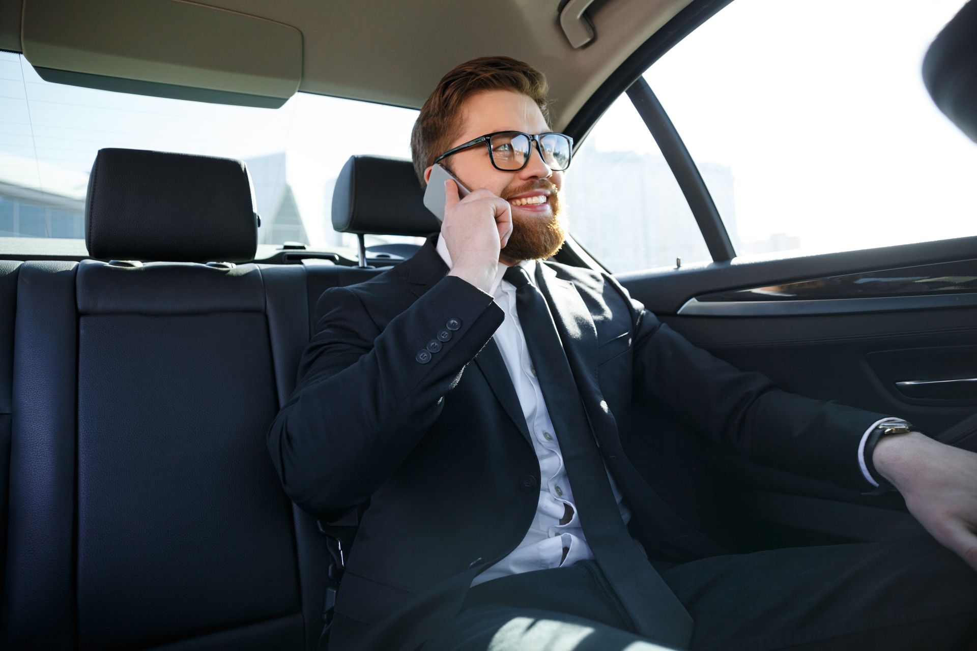 A smiling professional in a suit and glasses talks on a cell phone while sitting in the back seat of a car.