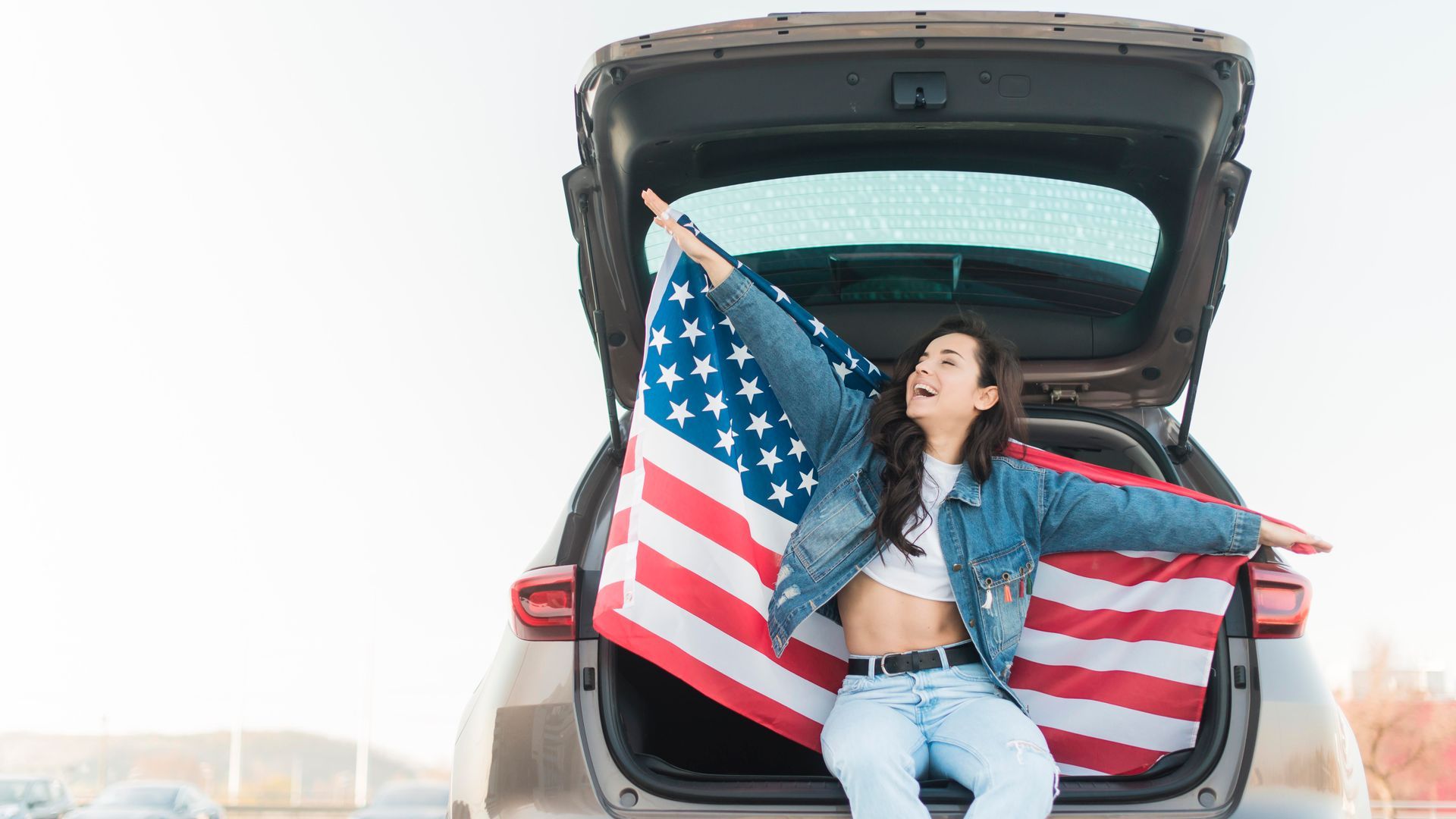 A person sits in the open trunk of a car, arms outstretched, holding a large American flag behind them.