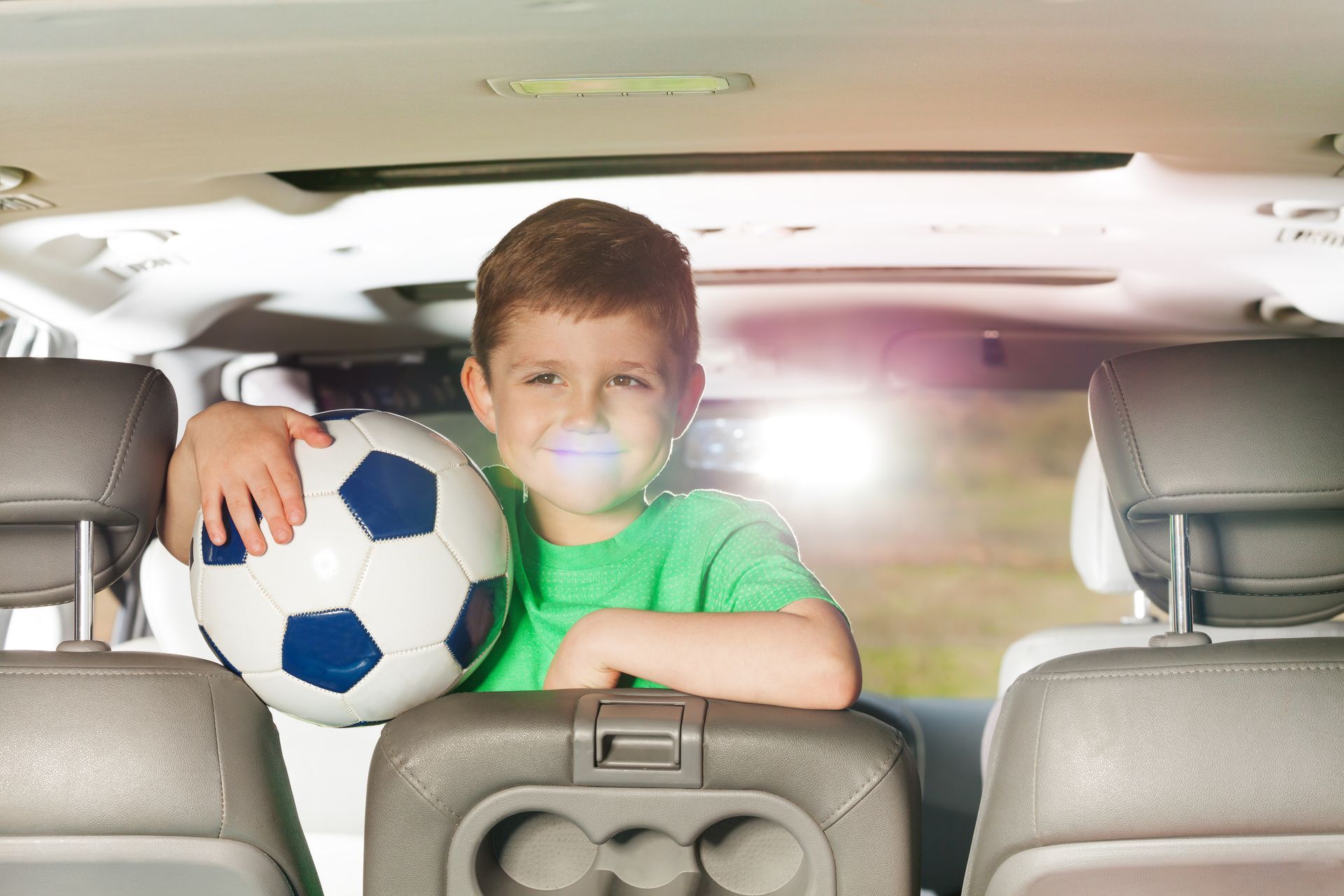 Boy in green shirt holding a soccer ball inside a car, smiling.