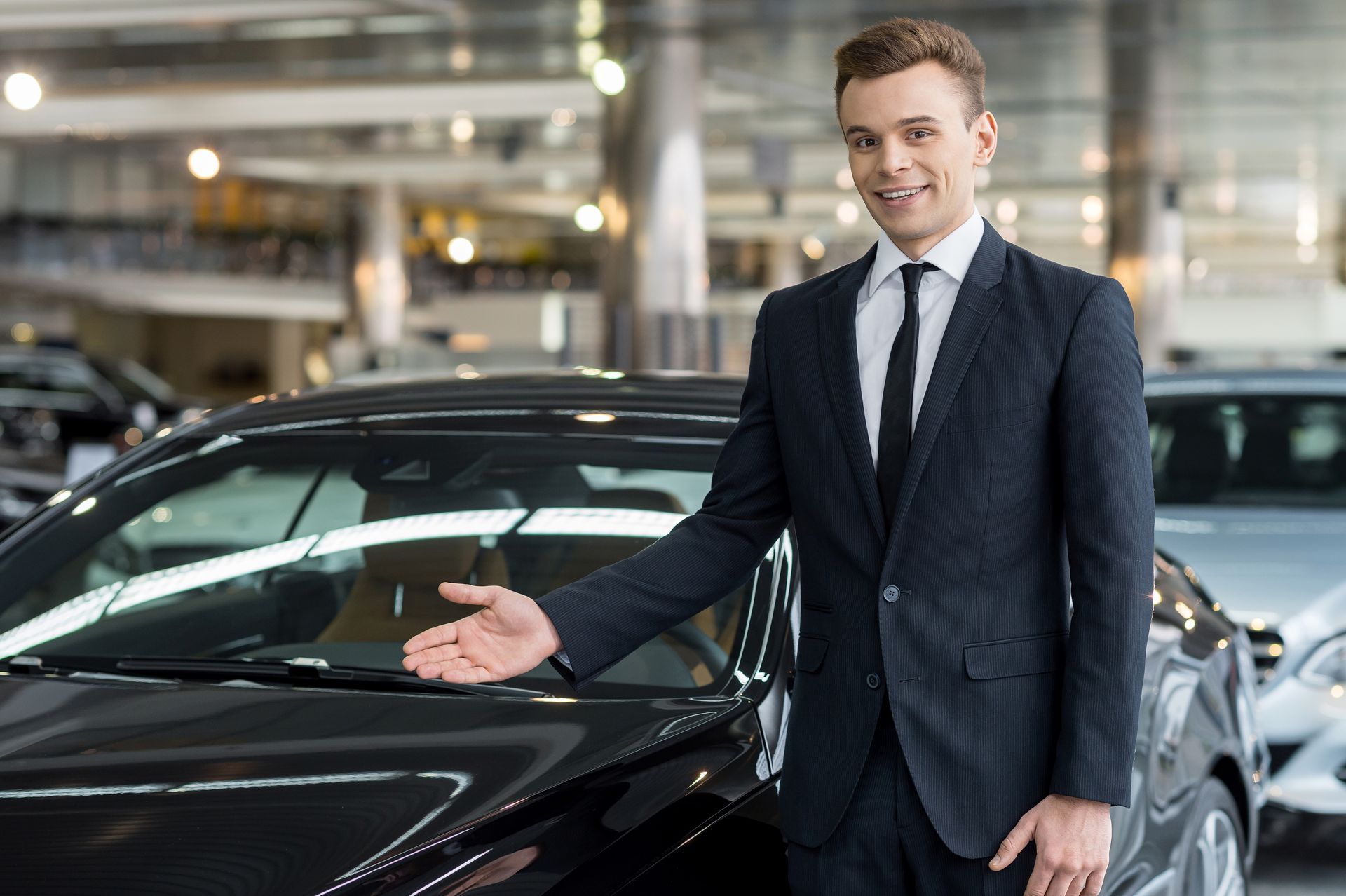 A man in a suit and tie is standing next to a black car in a showroom.