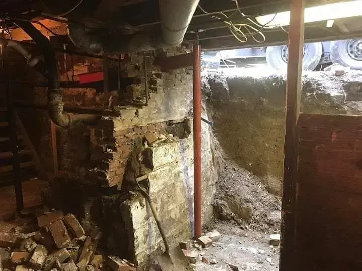 Dilapidated basement with exposed pipes, crumbling brick walls, and rubble piled near a damaged doorway