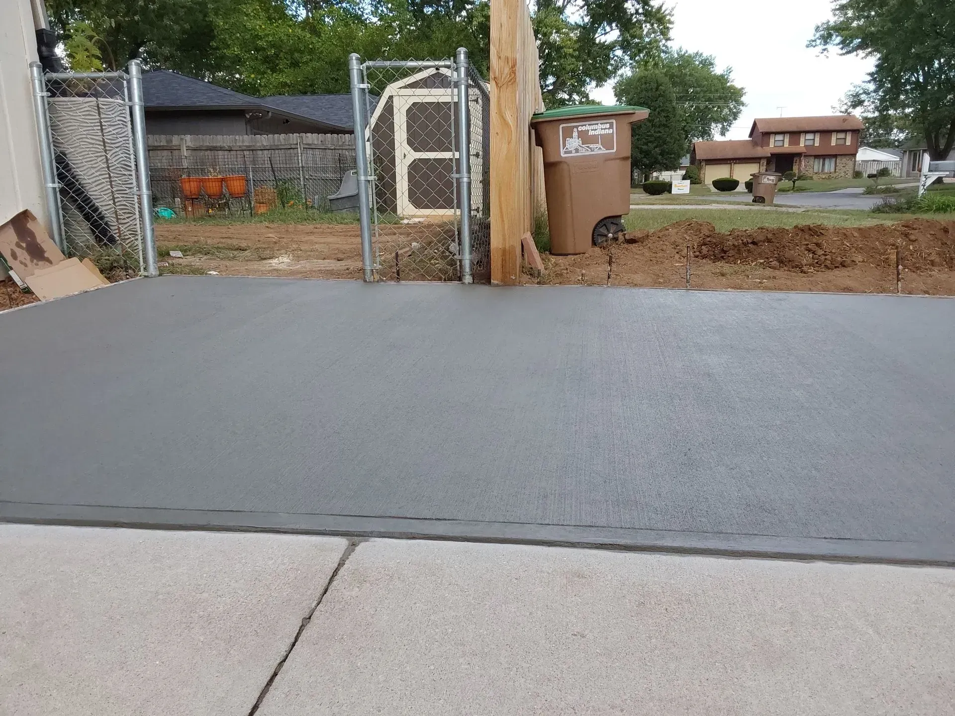 Freshly paved driveway with a gate, fence, and trash bin beside a dirt yard