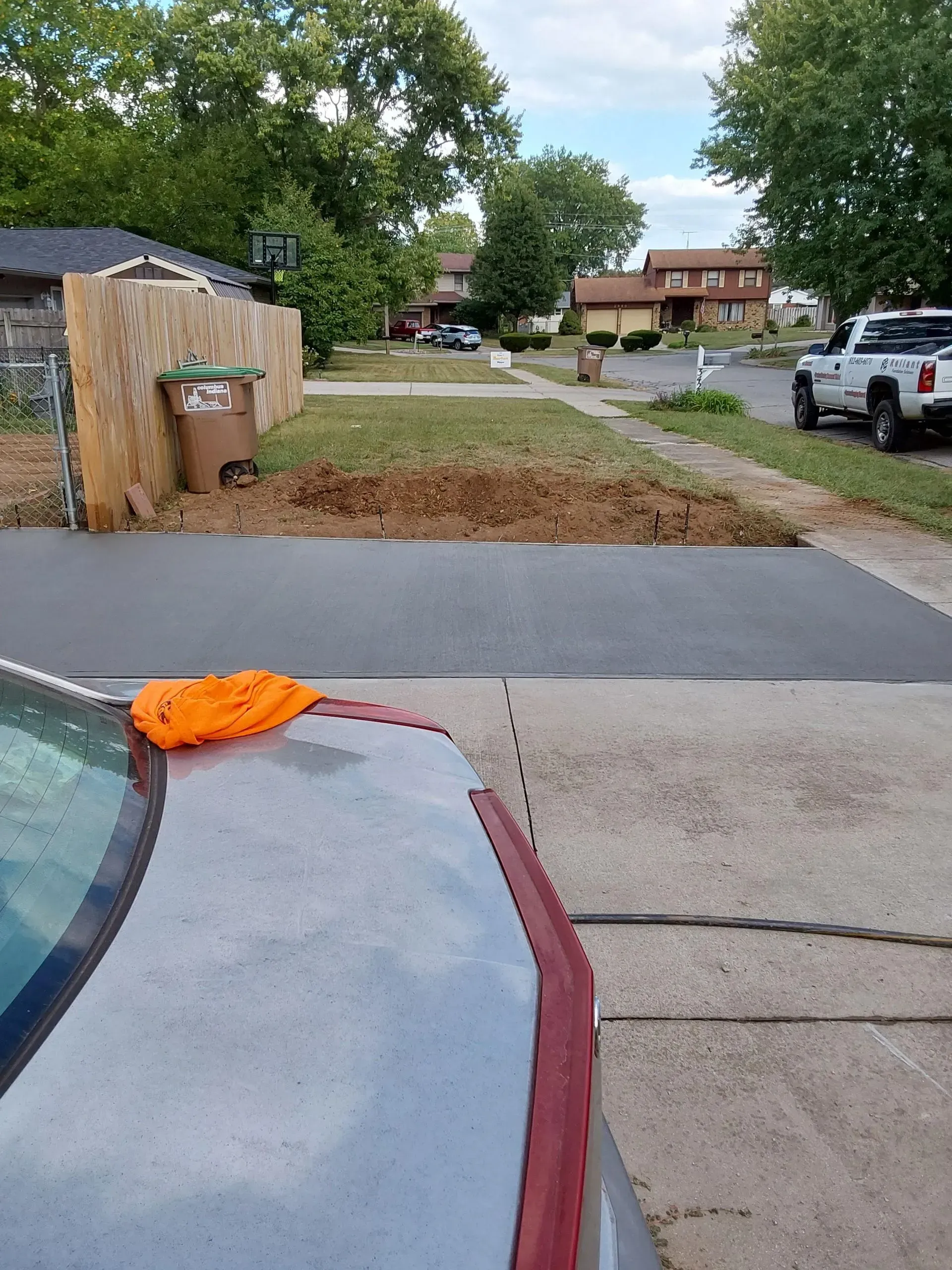 Driveway and dirt patch beside a house, with parked cars and a car hood in the foreground.