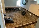 Water-damaged room with torn-up flooring and exposed subfloor, showing construction debris.