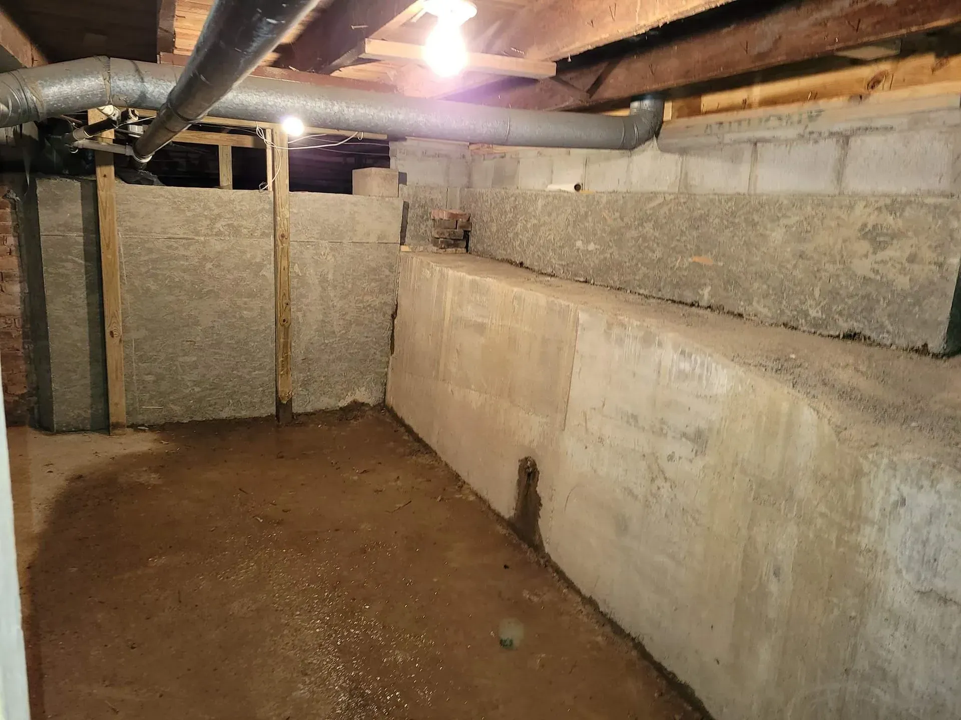 Basement crawlspace with dirt floor, concrete walls, exposed beams, and a single light bulb.