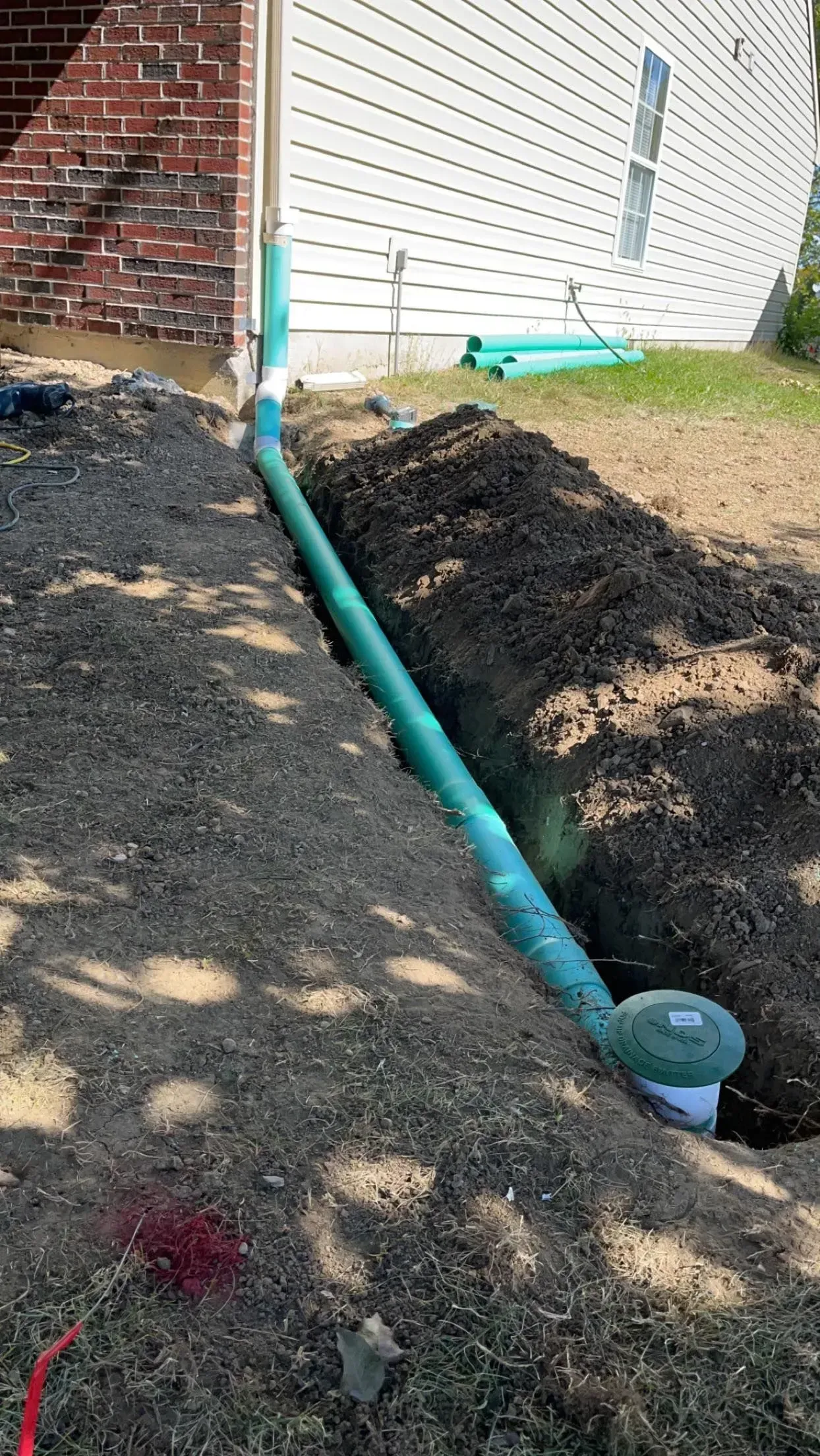 Green pipe running through a dirt trench beside a house foundation and brick wall.