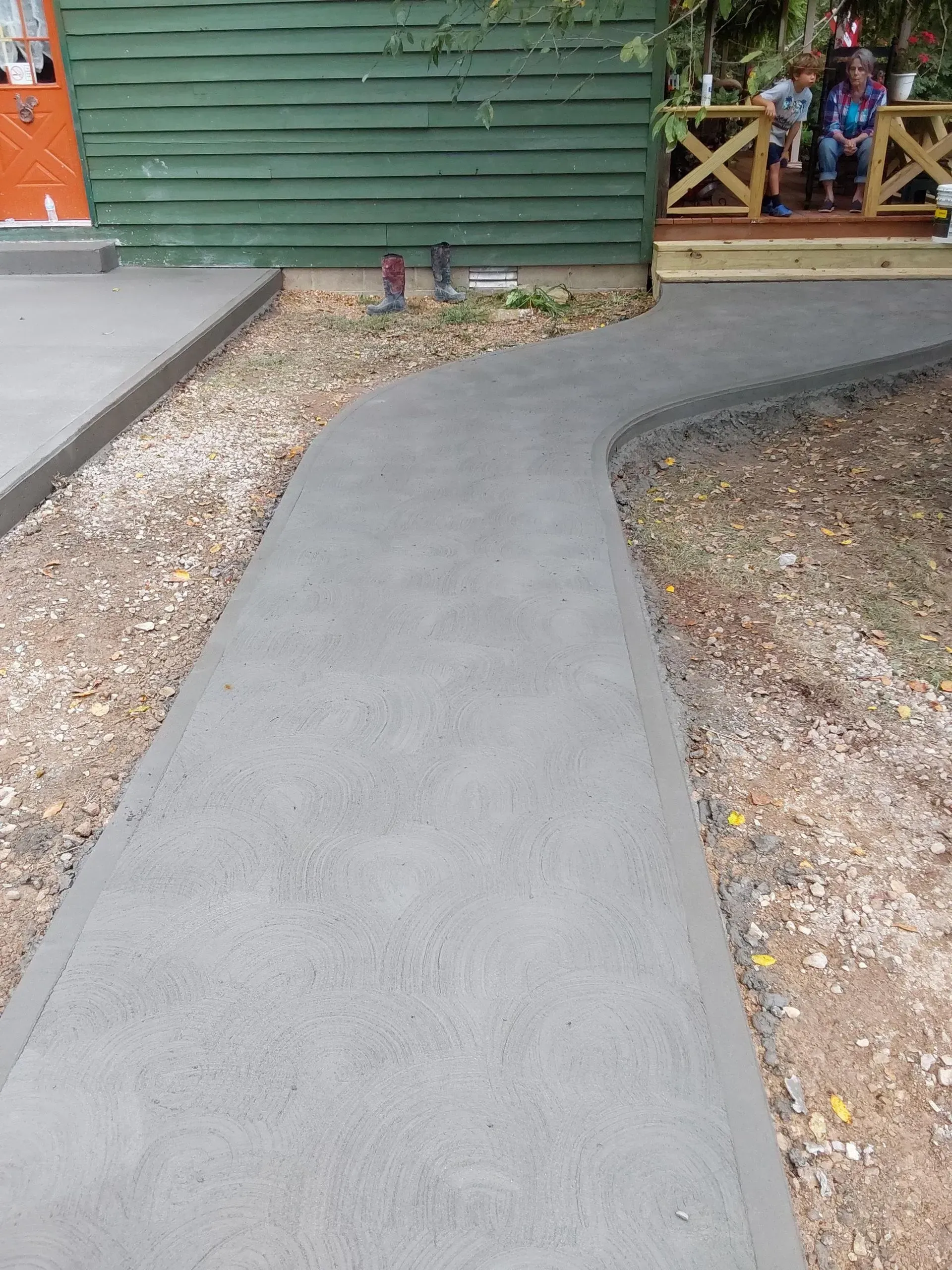 Concrete walkway curving past a green building toward a porch, bordered by gravel and dirt.