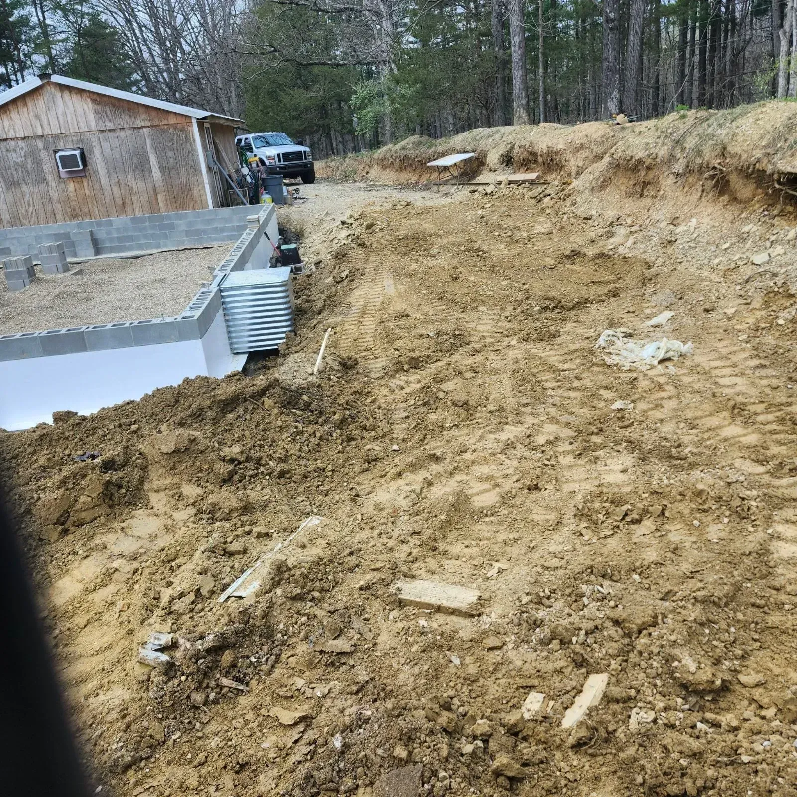 Cleared dirt slope beside a house foundation with a truck and wooded background