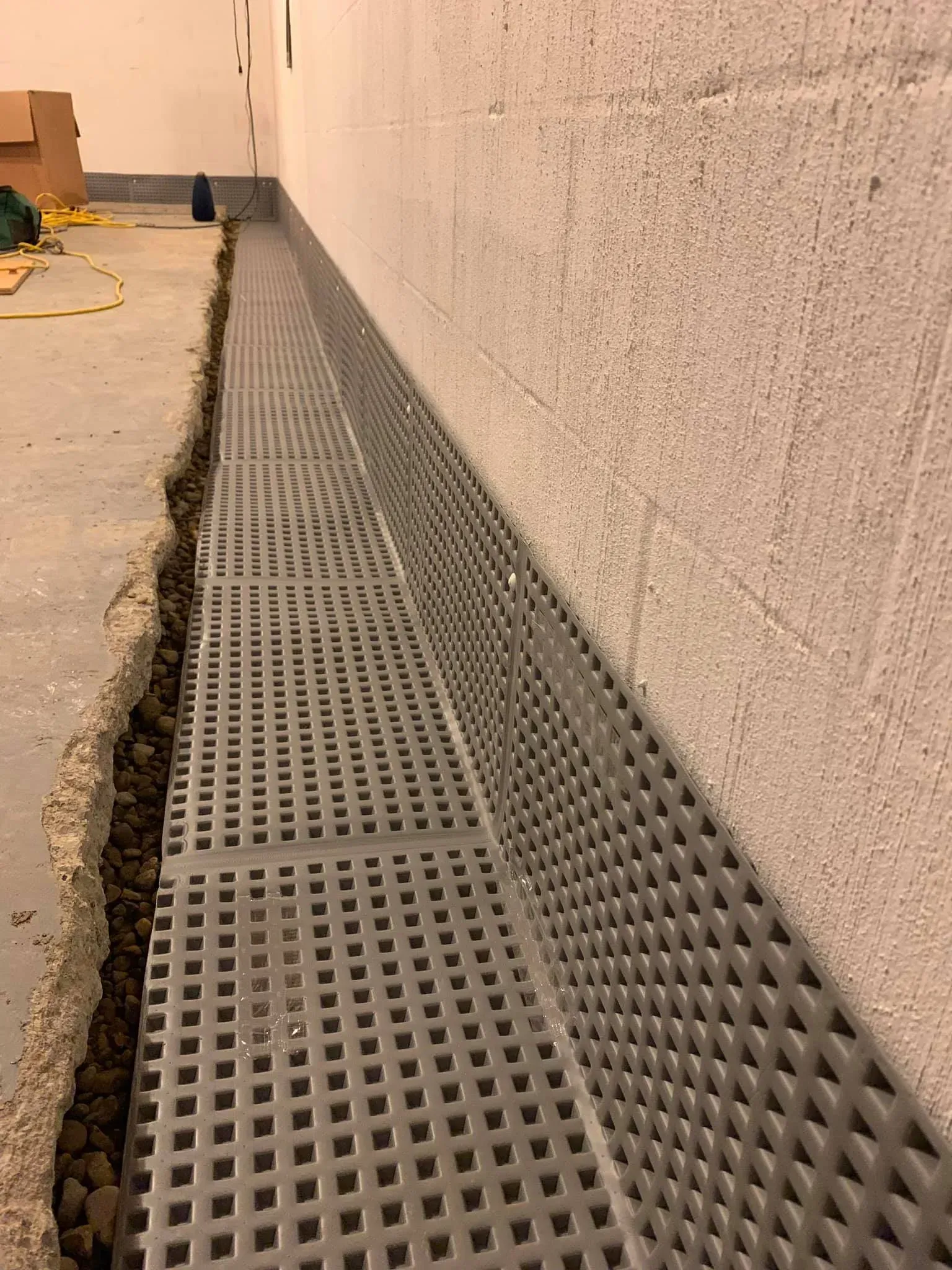 Perforated drainage grate along a wall beside a concrete walkway with a cracked edge