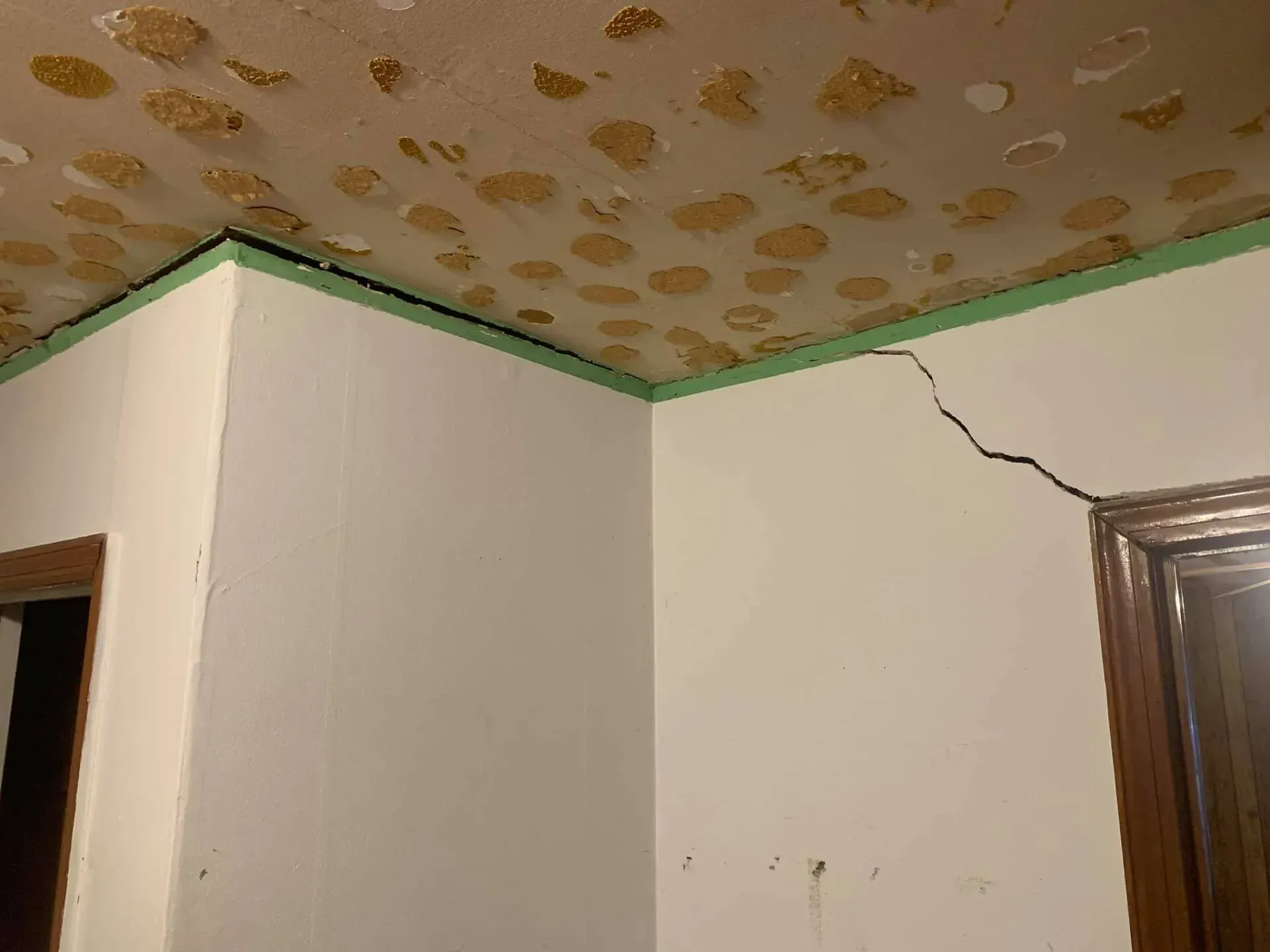 Damaged room corner with cracked white walls, green trim, and stained ceiling near a doorway