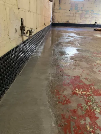 Wet concrete floor in a damaged, partially repaired room with a black drainage strip along the wall