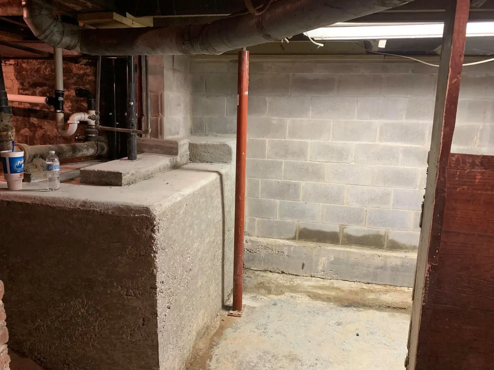 Basement crawlspace with exposed pipes, concrete block wall, and a utility tank in a dim area