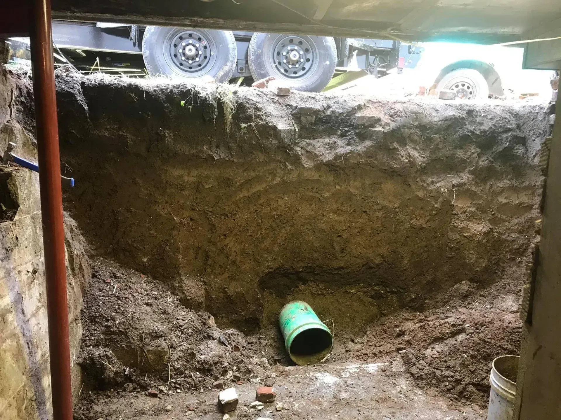 Excavated dirt pit with a green pipe opening, construction truck wheels visible above the edge.