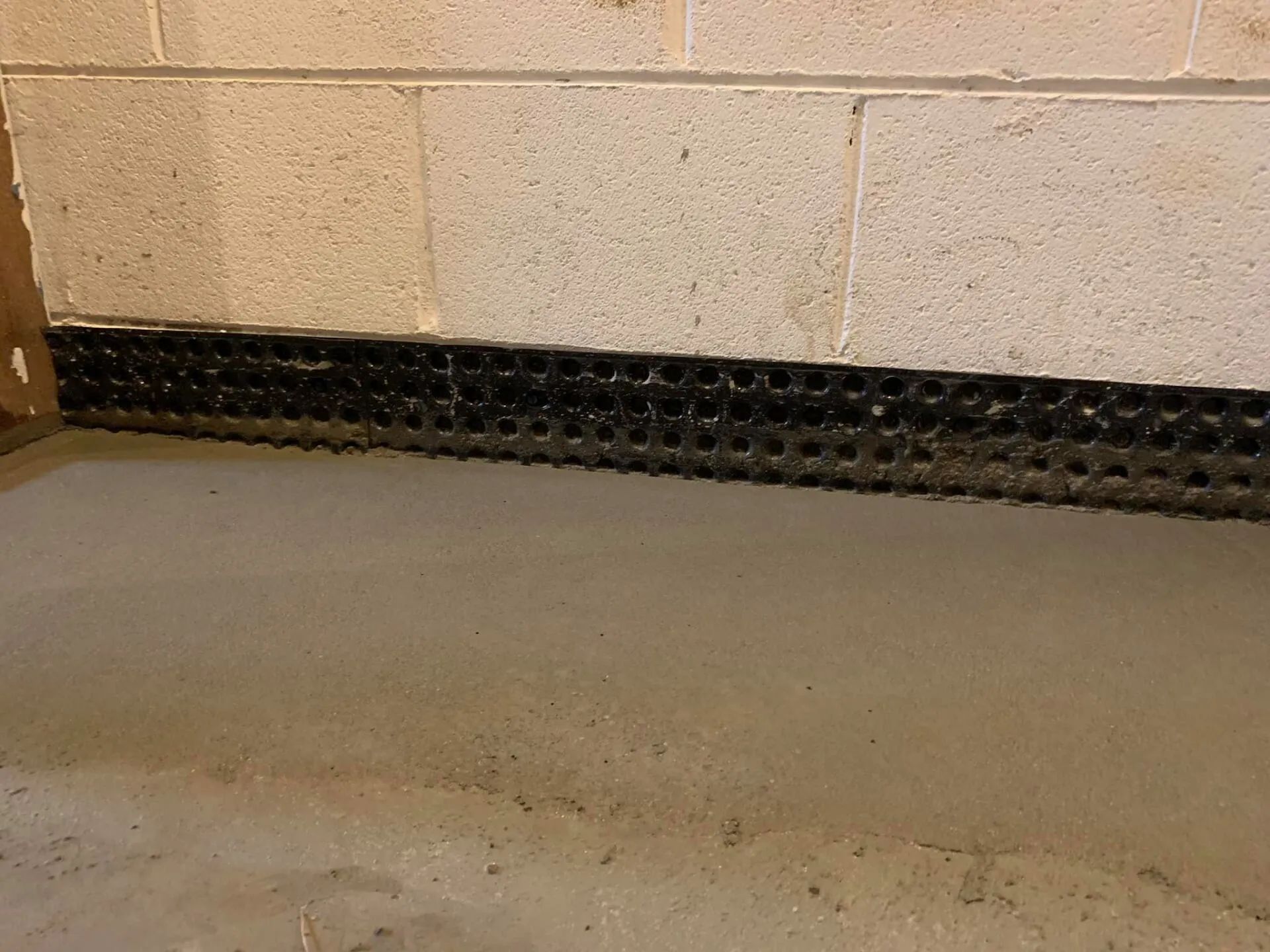 Basement wall with a black drainage system along the bottom. Beige concrete blocks above a gray concrete floor.