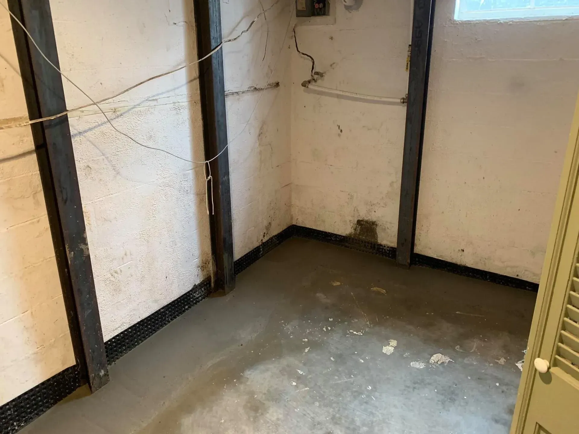 Empty corner of a basement room with gray floor, white walls, dark trim, and exposed pipes and cables.
