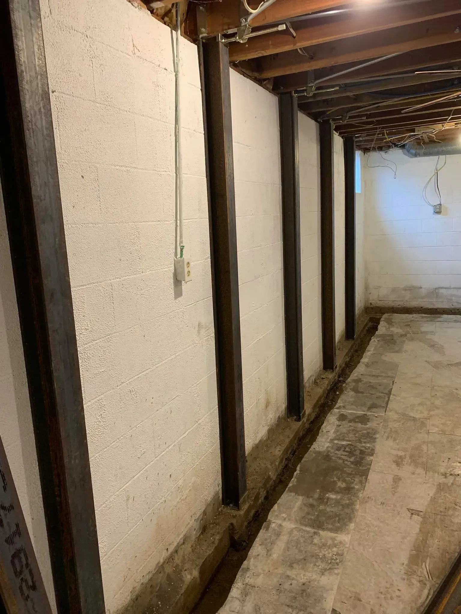 Narrow basement corridor with white walls, dark wooden beams, and a concrete floor.