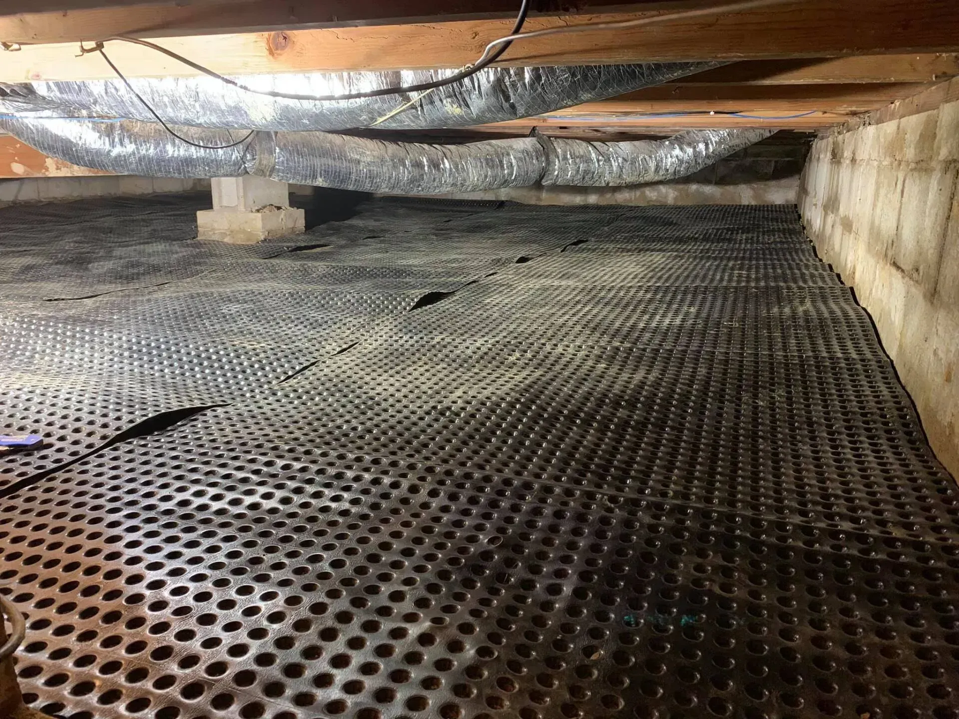Crawlspace with perforated black floor mat, exposed beams, ducting, and concrete foundation walls