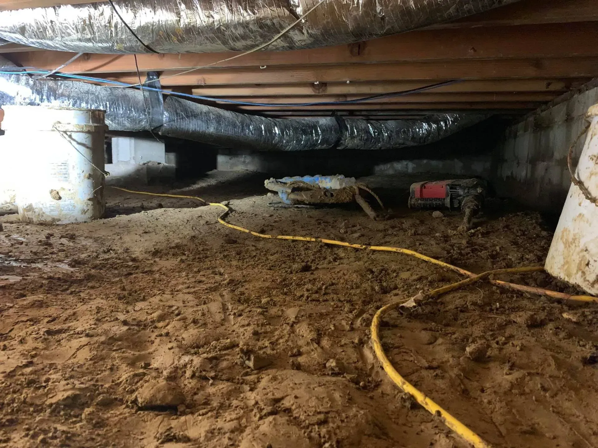 Mud-filled crawlspace under a house with exposed beams, pipes, and a yellow hose.
