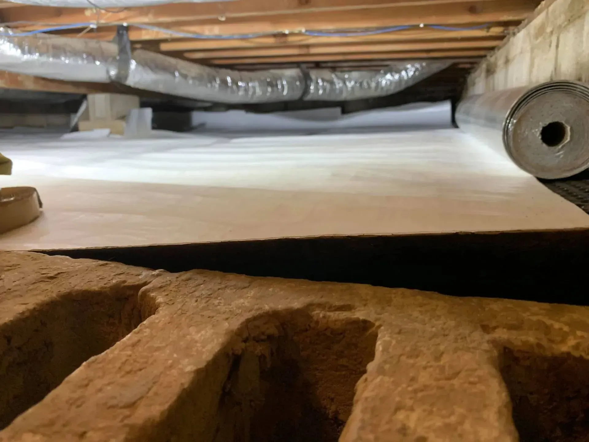 Inside a crawlspace, white insulation under wooden joists with a duct opening on the right.